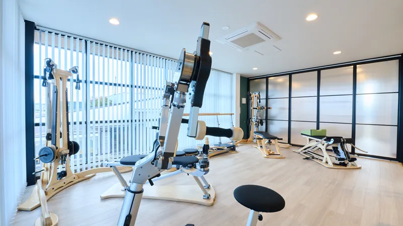 大きな窓から自然光が差し込む明るく清潔な室内に、木製の専用トレーニング・リハビリマシンが複数設置されている様子
