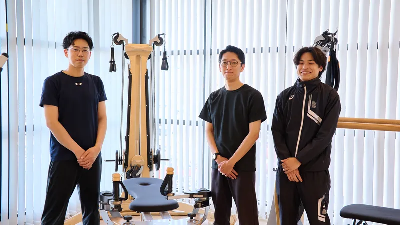 明るい窓際にある木製の専用マシンの前に並んで立つ、スポーティーな服装の3人の男性トレーナー。手を前に組み、穏やかな表情でカメラに視線を向けている様子